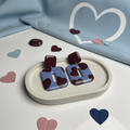 Paar moderne Statement-Ohrringe in Pastellblau mit bordeauxfarbenem Herzmotiv und glänzender Oberfläche, arrangiert auf einer hellen ovalen Ablage, umgeben von dekorativen Papierherzen auf hellem Hintergrund.